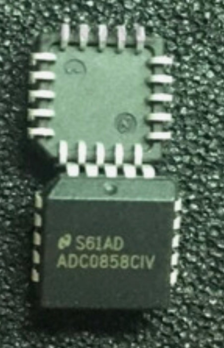 A black integrated circuit chip with white printed text reading 'S61AD ADC0858CIV' and multiple white pins around the edges, photographed on a dark textured surface.