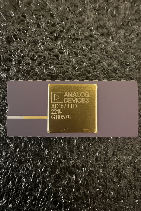 Analog Devices AD1674TD analog-to-digital converter chip in its original dark gray and gold packaging, resting on a textured black surface.